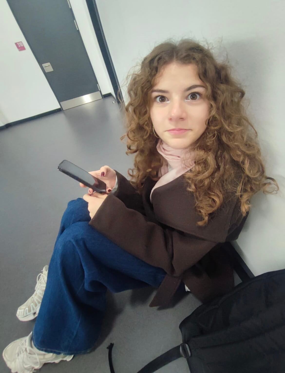 Das Bild zeigt Lara, wie sie sitzend ein Handy hält und in die Kamera schaut.